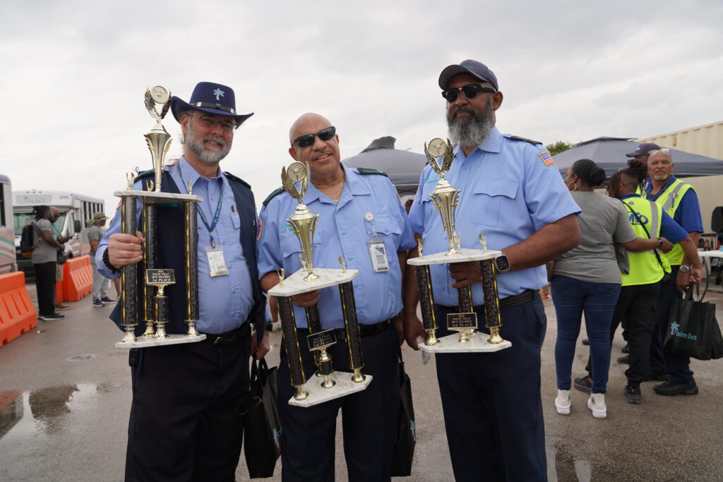 Three uniformed men stand outdoors holding large trophies, with other people and event tents visible in the background.