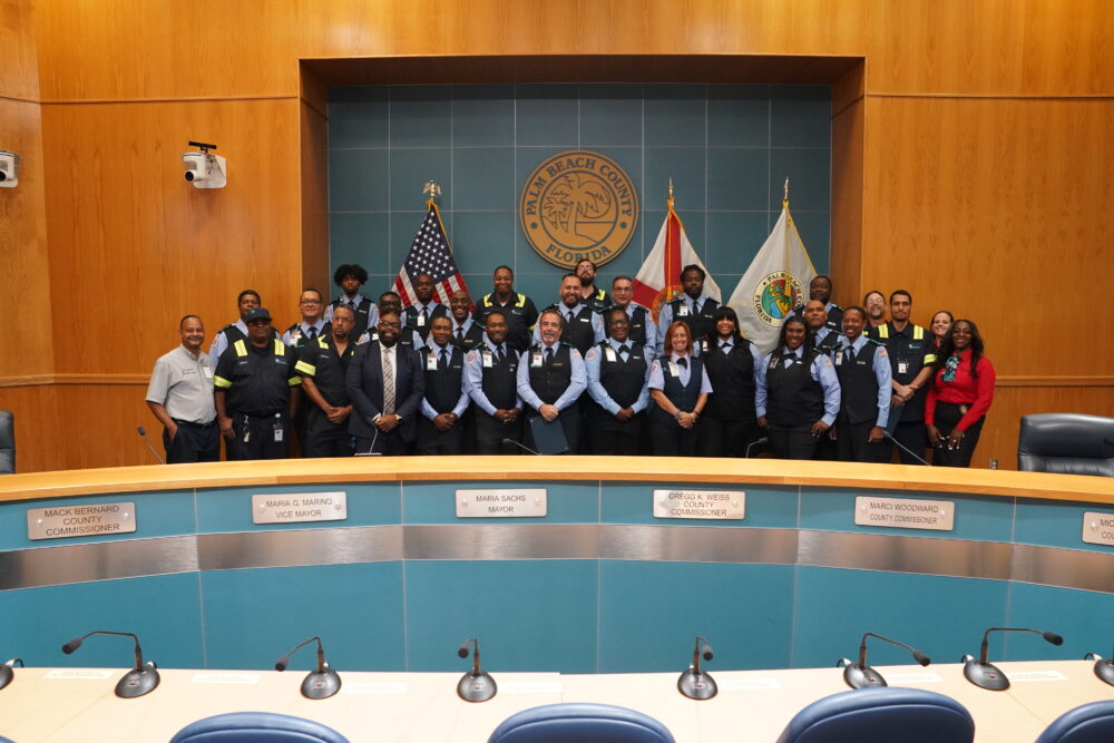 A large group of uniformed employees poses together in a government meeting room in front of flags and the Palm Beach County, Florida seal.