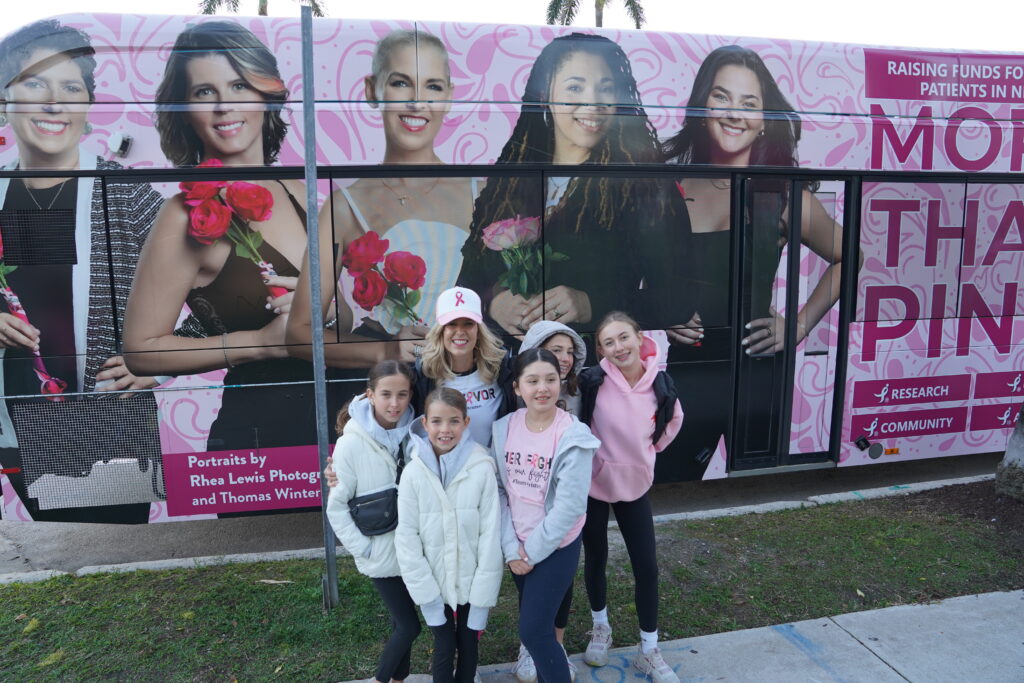 Five people, four children and one adult, pose and smile in front of a pink bus covered with portraits of women and breast cancer awareness graphics.