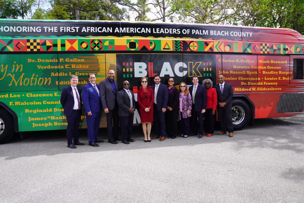 A group of people in formal attire stand in front of a colorful bus honoring the first African American leaders of Palm Beach County.