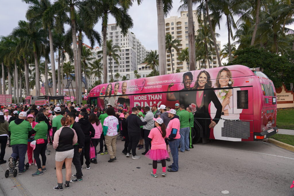 A crowd of people, many in pink clothing, gathers near a large pink bus with women's images and the phrase "More Than Pink" on a city street lined with palm trees and tall buildings.
