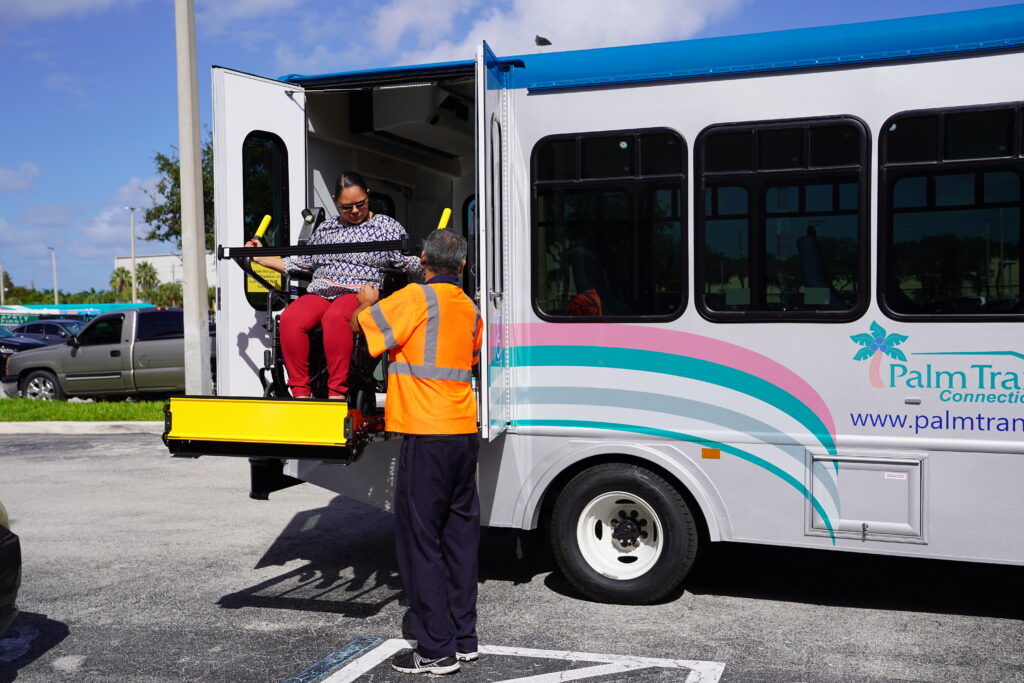 A person in a wheelchair is being assisted onto a Palm Tran Connection bus using a wheelchair lift while an attendant in an orange vest supervises.
