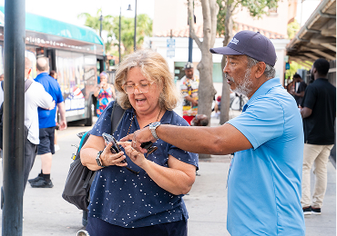 A man in a blue uniform helps a woman with her smartphone at a bus stop, with a bus and other people visible in the background.