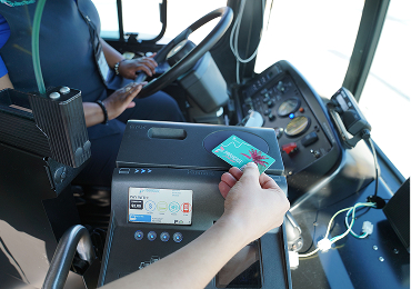 A person taps a transit card on a fare reader inside a bus while the driver sits at the wheel.