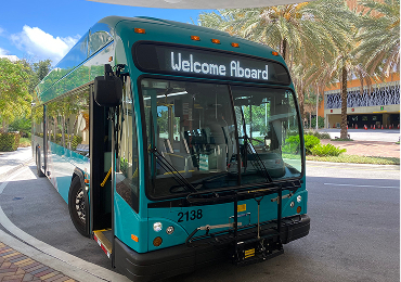A teal and black city bus with "Welcome Aboard" displayed on its sign is parked at a curbside under palm trees.