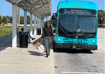 A woman walks away from a blue city bus labeled "3 Northbound via Military to Gardens Mall" at a bus stop on a sunny day.