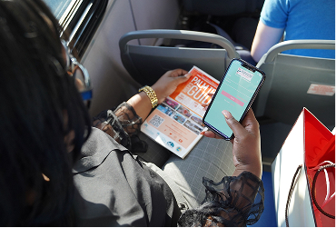 A person sitting on a bus holds a pamphlet in one hand and a smartphone in the other, with another passenger seated ahead.