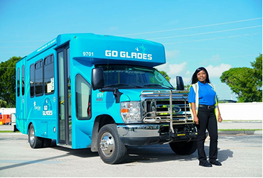 A woman in uniform stands next to a blue GO GLADES shuttle bus parked on a sunny day.