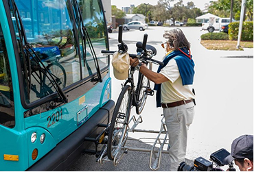 A person places a bicycle onto the front bike rack of a blue public bus parked at a curb on a sunny day.