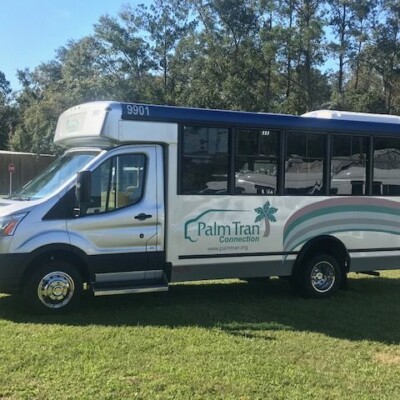 White Palm Tran Connexion shuttle bus parked on grass, displaying the service logo and number 9901, with trees and buildings in the background.