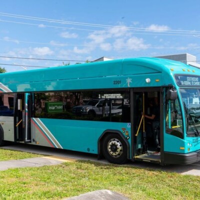 A teal public transit bus is stopped at a curb on a sunny day, with its door open and a white van parked behind it.