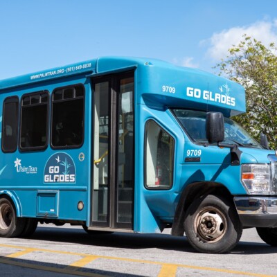 A blue Palm Tran Go Glades shuttle bus is parked near a sidewalk on a sunny day, with trees and a streetlamp in the background.