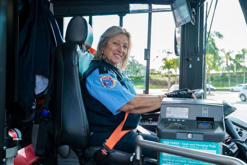 A smiling bus driver in uniform sits behind the wheel, with a view of trees and a street through the windshield. The scene conveys a sense of friendliness and professionalism.