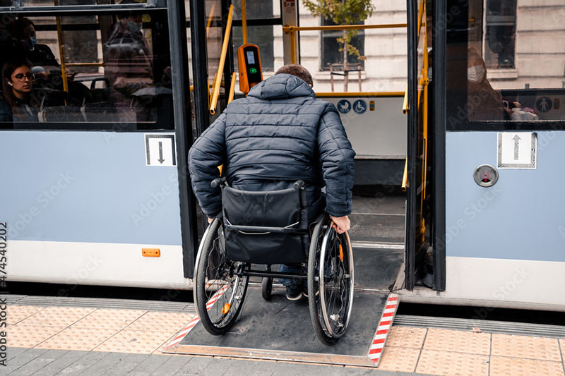 A person in a wheelchair boards a bus using a ramp, wearing a dark jacket. The scene conveys accessibility and independence in urban transit.