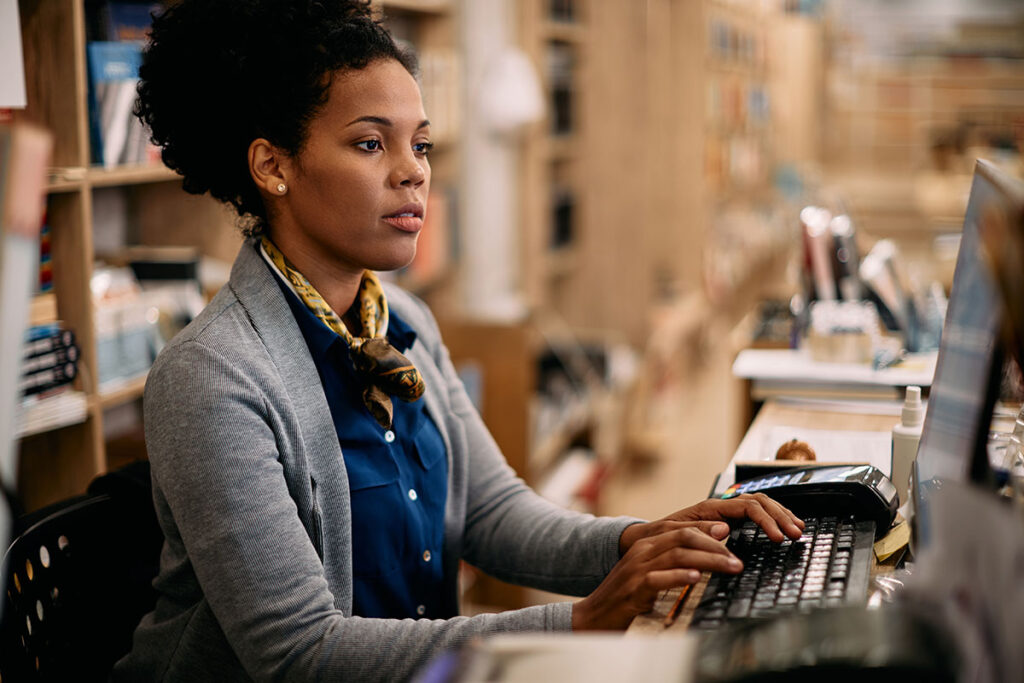 A focused woman in a blue shirt and grey cardigan works at a computer in an office filled with shelves. The atmosphere is professional and attentive.