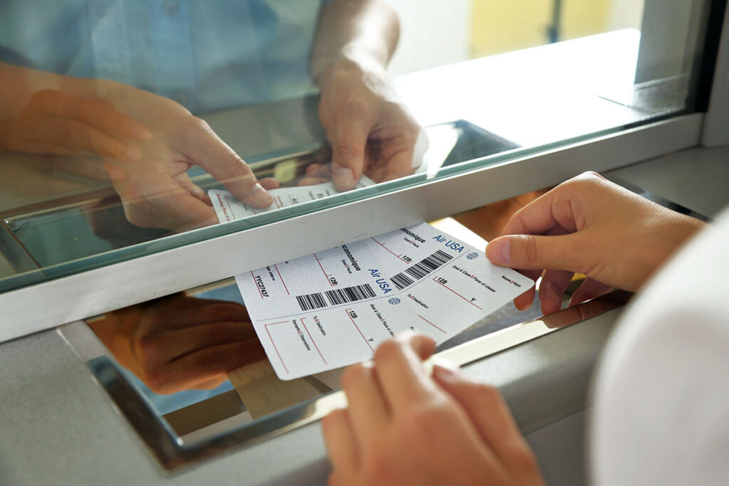 Hands exchanging airline tickets through a counter window, with focus on the tickets displaying flight details. Professional, travel-related setting.