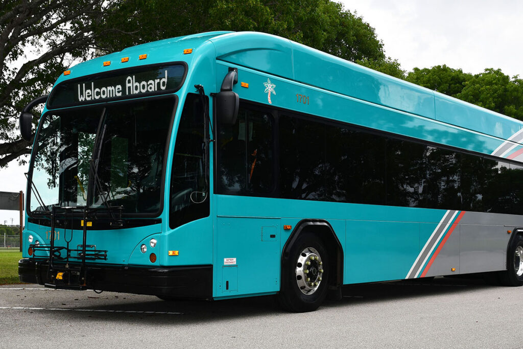 Turquoise city bus on a road, displaying "Welcome Aboard" on the front. The bus is surrounded by green trees, creating a welcoming and friendly scene.