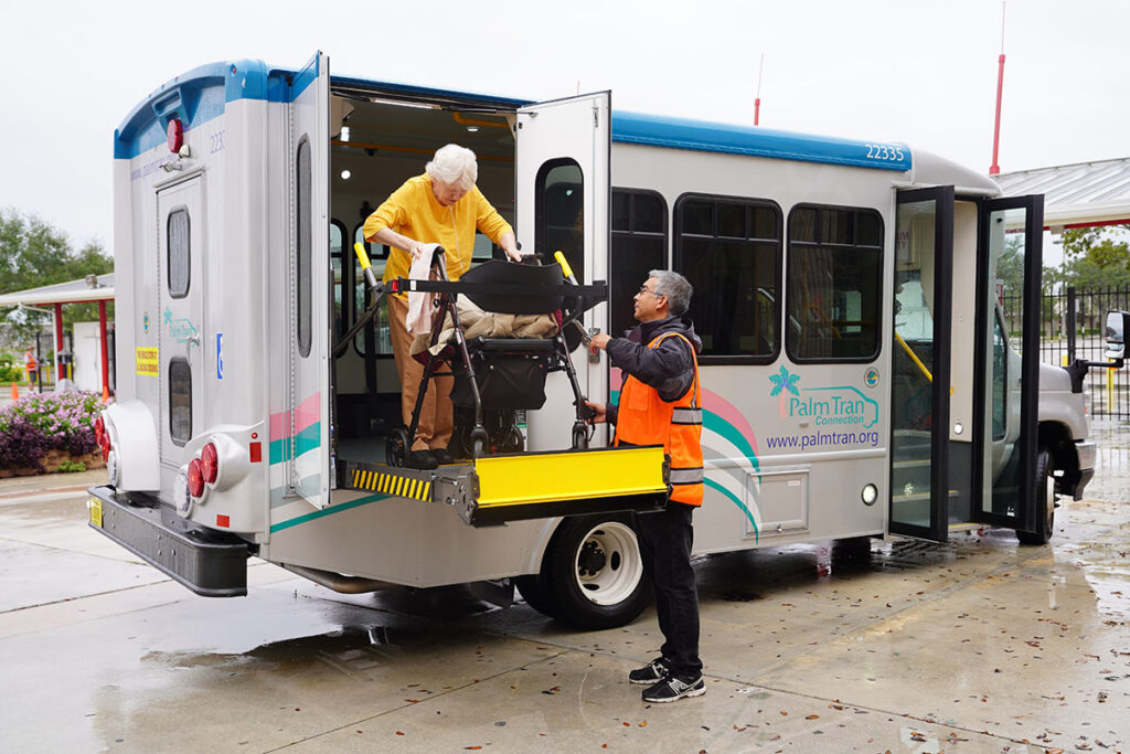 A senior woman with a walker uses a lift to board a Palm Tran Connects van. A man in an orange vest assists. The scene is calm and supportive.