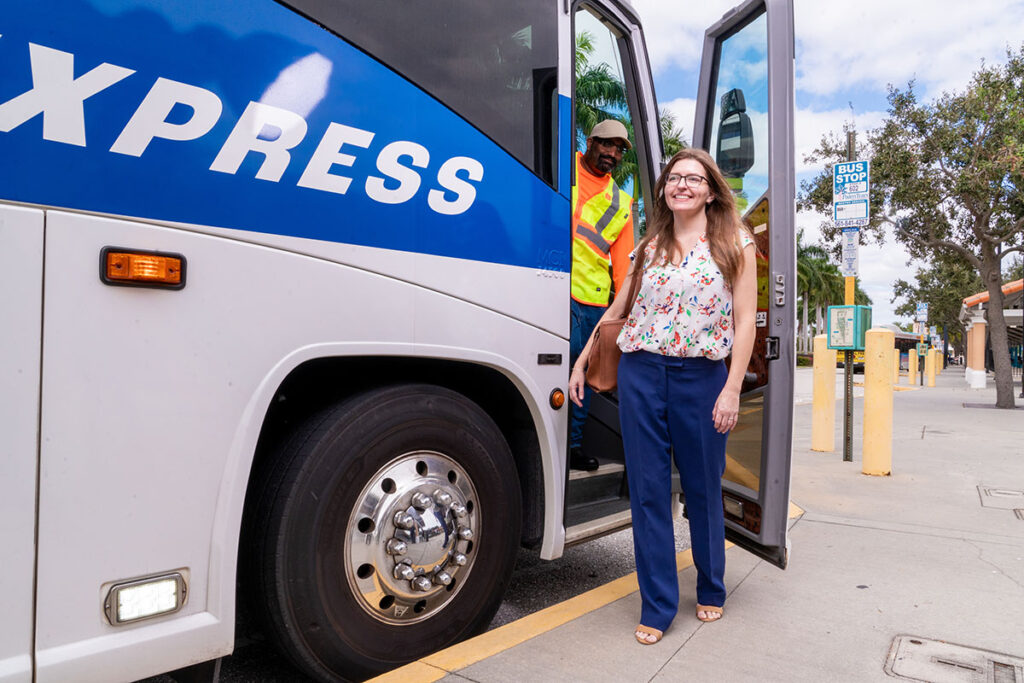 Smiling woman steps off a blue and white express bus, assisted by a bus driver in a safety vest. Sunny day at a bus stop with trees and signs.