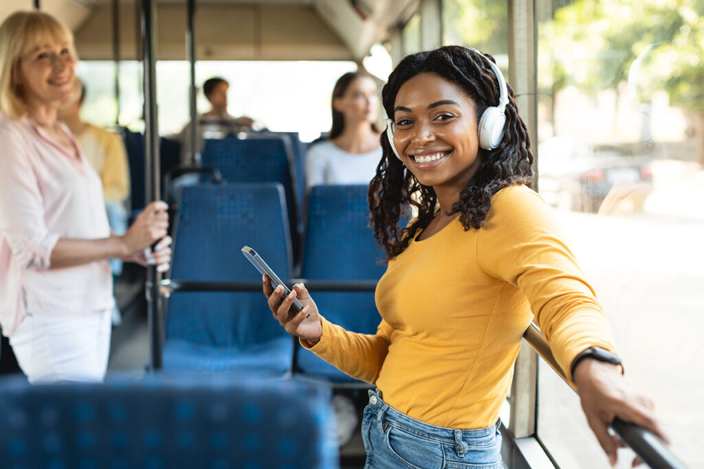 A woman in a yellow top listens to music on headphones and smiles while holding a phone on a bus. Other passengers are seated in the background.
