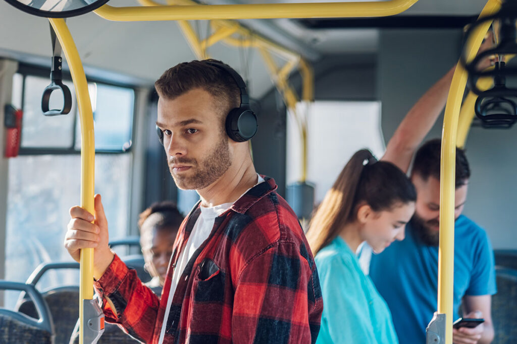 Man in a red plaid shirt with headphones stands on a bus, holding a yellow rail. Other passengers are smiling, suggesting a relaxed mood.