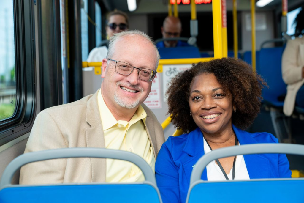 A smiling man and woman sit together on a bus. The man wears glasses and a beige suit, while the woman is in a bright blue blazer. They convey warmth and friendliness.