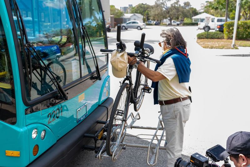 A person places a bicycle onto the front bike rack of a blue public bus parked at a curb on a sunny day.