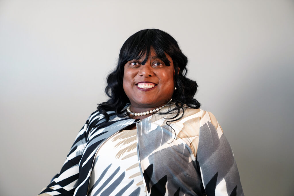 A person with long black hair, wearing a patterned jacket and pearl necklace, smiles at the camera against a plain light-colored background.