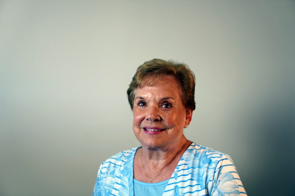 An older woman with short light brown hair, wearing a light blue patterned shirt, smiles in front of a plain, light-colored background.
