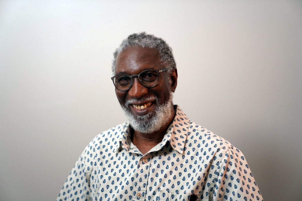 An older man with gray hair, a beard, and glasses smiles while wearing a light-colored patterned shirt against a plain, light background.