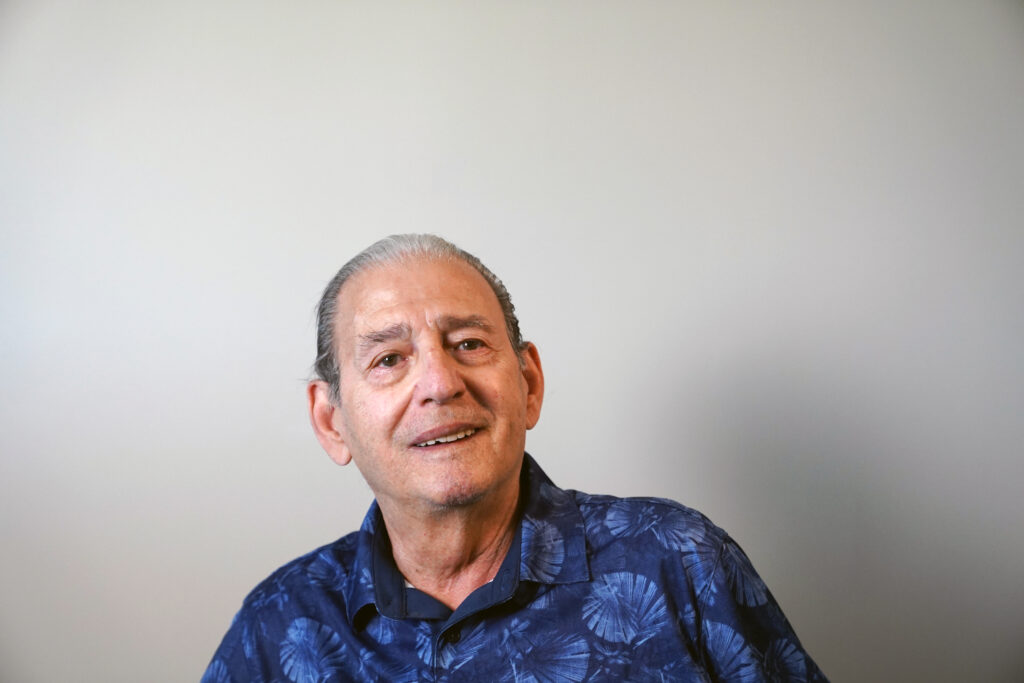 An older man with gray hair, wearing a blue patterned shirt, sits and looks slightly upward against a plain light-colored background.