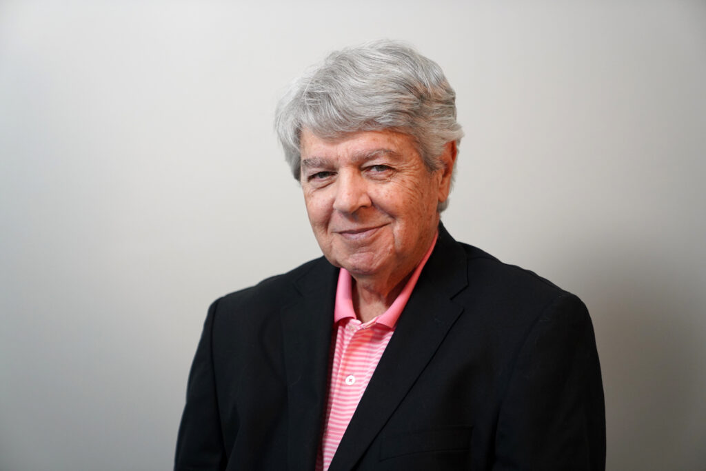 An older man with gray hair wearing a black blazer over a pink collared shirt stands in front of a plain light background.