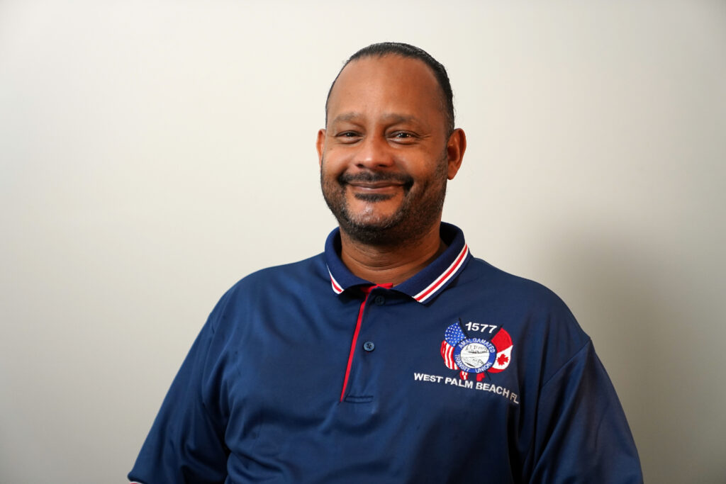 A man wearing a blue collared shirt with West Palm Beach, FL and American and Canadian flags on it stands against a plain light background, smiling at the camera.