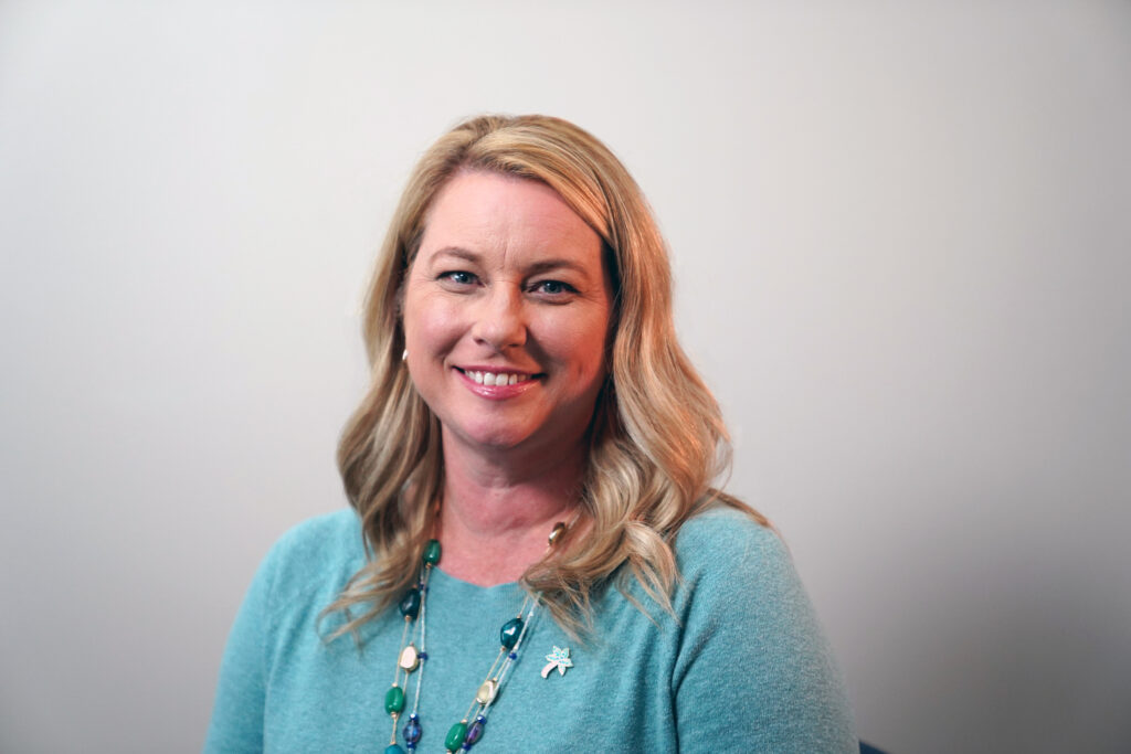 A woman with blonde hair, wearing a light blue sweater and a colorful beaded necklace, smiles at the camera against a plain background.