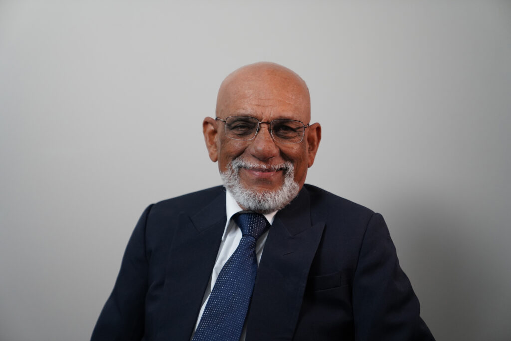 An older man with a bald head, white beard, and glasses, wearing a navy suit and blue tie, sits and smiles in front of a plain light gray background.