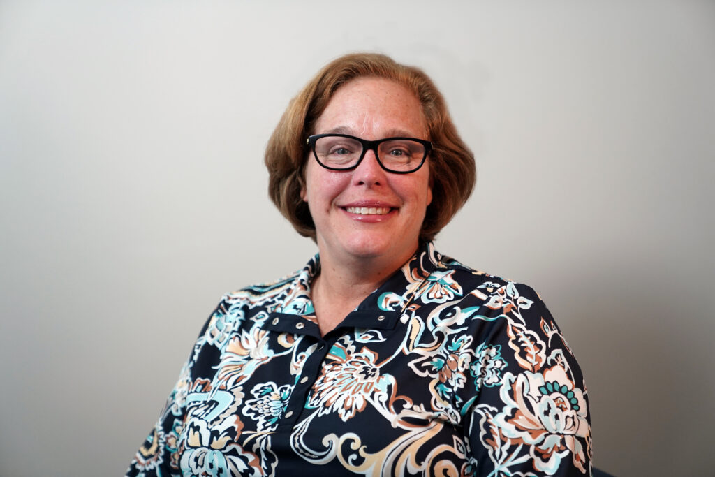 A person with short light brown hair and glasses, wearing a patterned shirt, sits and smiles in front of a plain light-colored background.