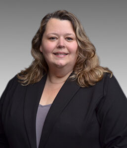 A woman with wavy, shoulder-length hair wearing a black blazer and a light gray top, posing against a plain gray background.
