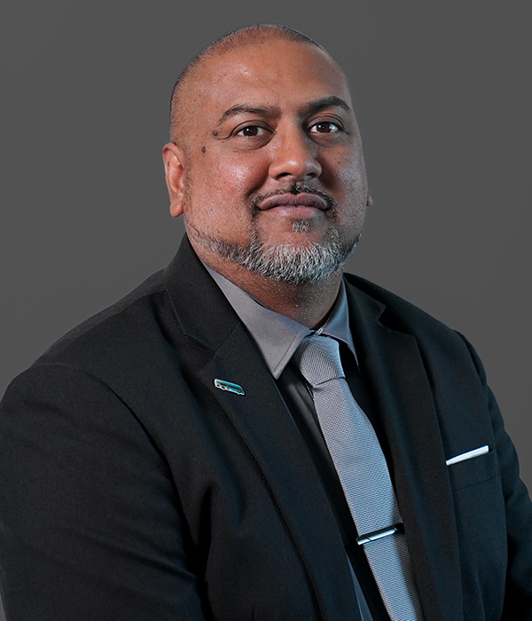 A man with a beard in a dark suit, light gray tie, and light blue shirt poses against a plain gray background.