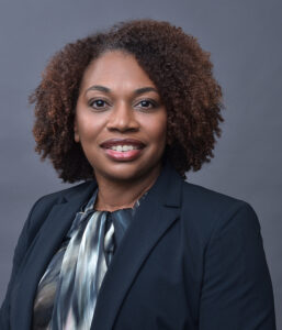 A woman with curly hair wearing a dark blazer and patterned blouse smiles at the camera against a plain gray background.