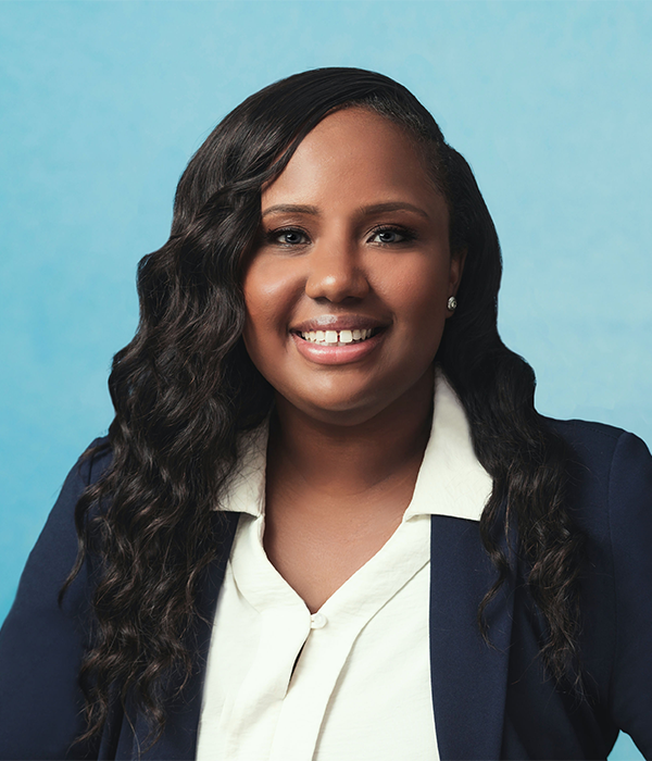 A woman with long, wavy hair wearing a navy blazer and white blouse smiles at the camera against a gray background.