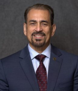 A man in a dark suit, light shirt, and patterned tie is smiling at the camera against a plain, dark background.