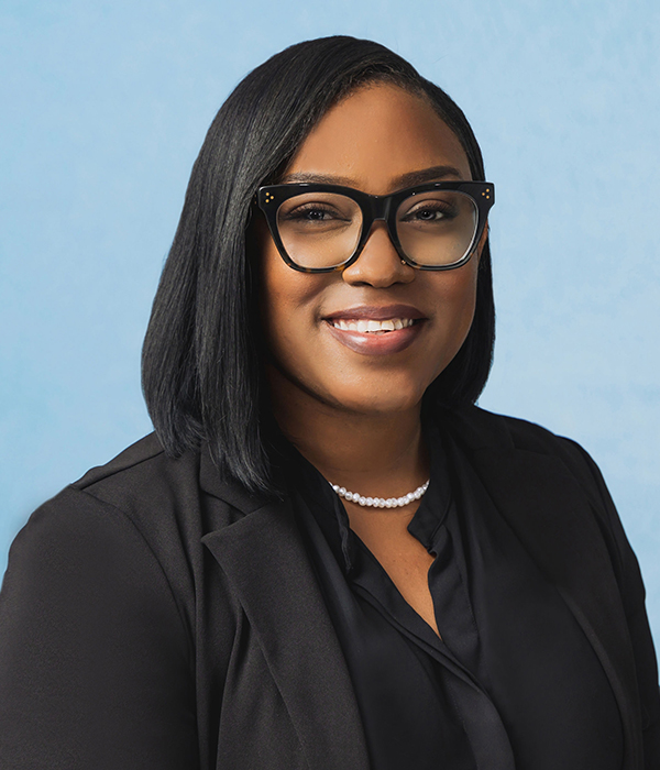 A woman wearing glasses, a black blazer, a black blouse, and a pearl necklace smiles at the camera against a light blue background.
