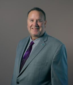 A man in a gray suit, lavender shirt, and purple tie stands against a plain background, smiling at the camera.