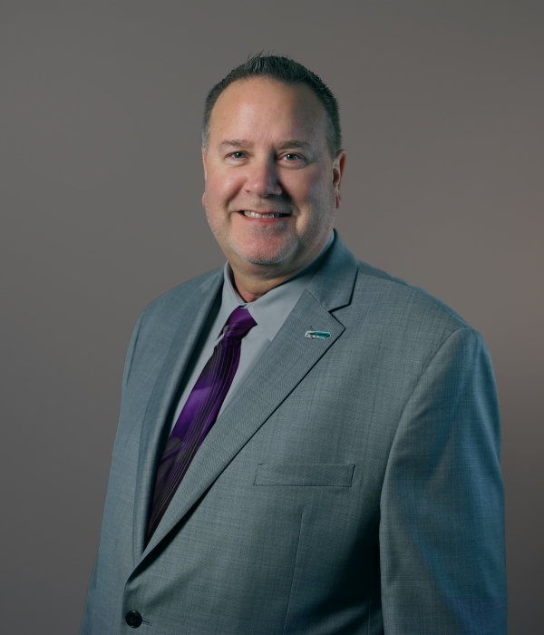 A man in a gray suit, lavender shirt, and purple tie stands against a plain background, smiling at the camera.
