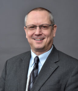 A middle-aged man wearing glasses, a gray suit, light blue shirt, and striped tie poses for a professional headshot against a plain gray background.