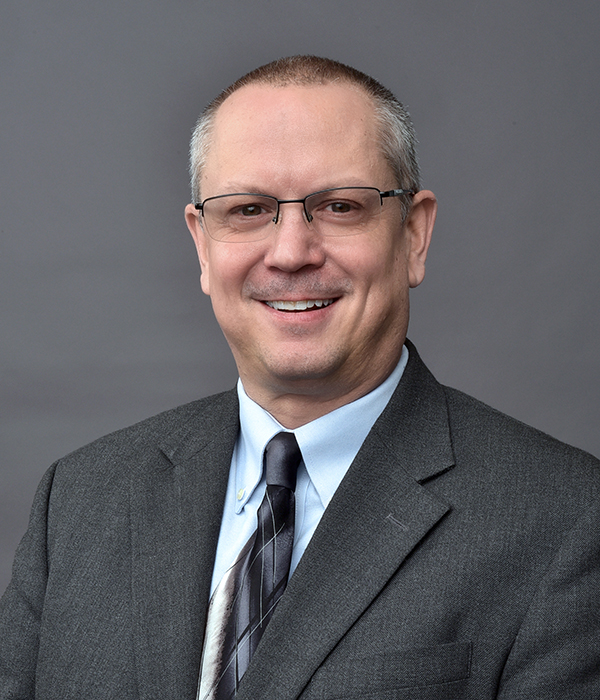 A middle-aged man wearing glasses, a gray suit, light blue shirt, and striped tie poses for a professional headshot against a plain gray background.