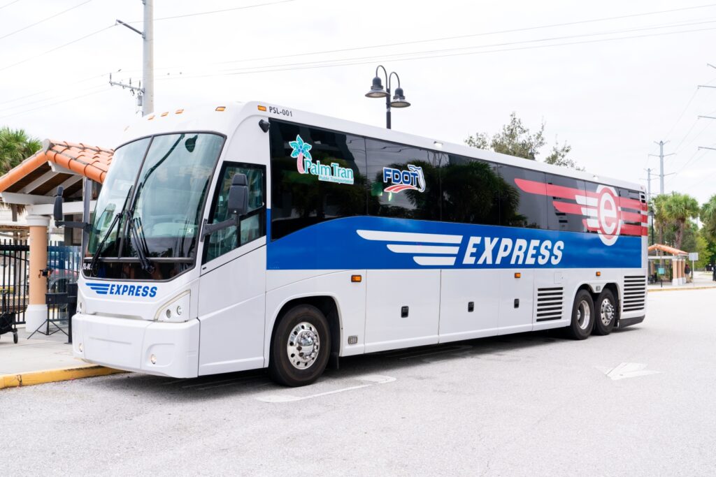 A large blue, white, and black express bus is parked at a bus stop, displaying the Palm Tran and FDOT logos on its side.