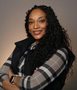 Woman with long curly hair wearing a black vest and plaid shirt poses with arms crossed, smiling against a plain background.