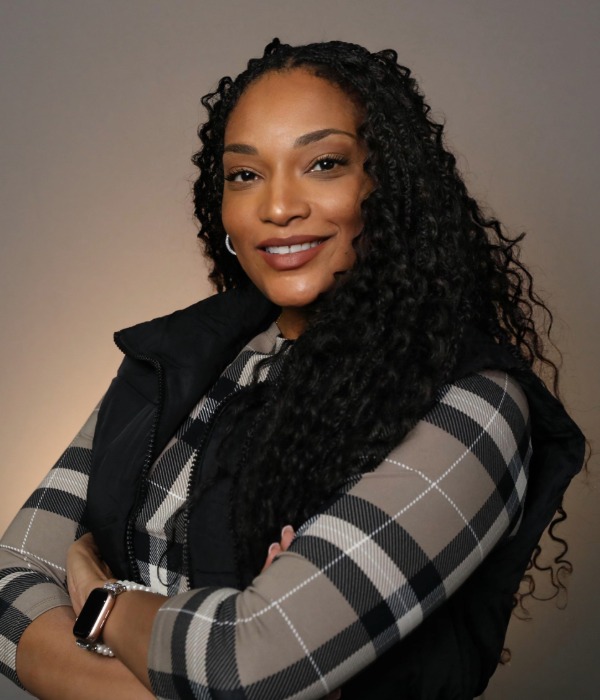 Woman with long curly hair wearing a black vest and plaid shirt poses with arms crossed, smiling against a plain background.