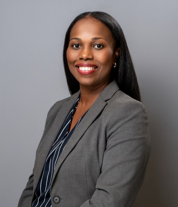 A woman in a gray blazer and striped blouse smiles at the camera against a plain gray background.